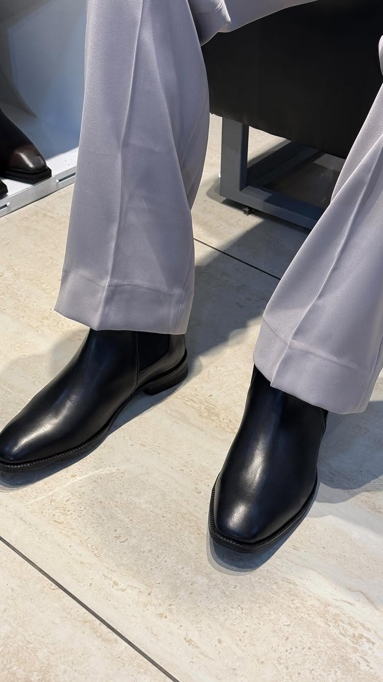 Black leather Chelsea boots paired with light grey tailored trousers on tiled flooring.