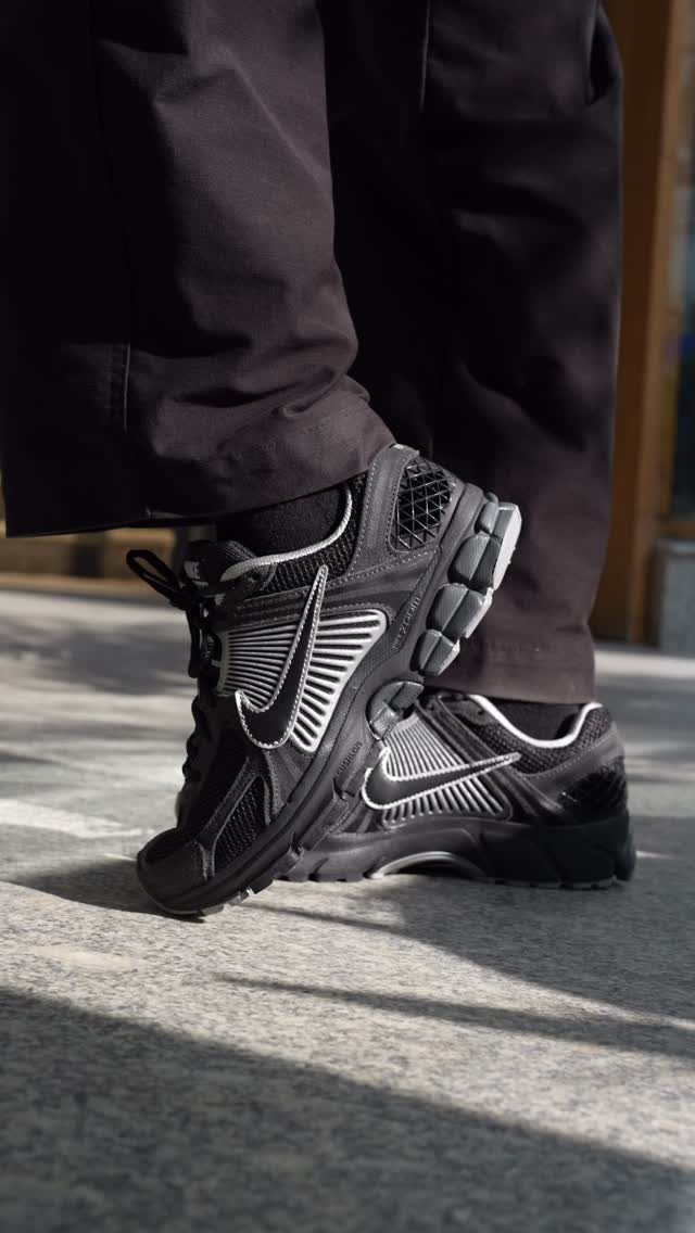 Black Nike Zoom Vomero 5 sneakers worn with black trousers on stone pavement.