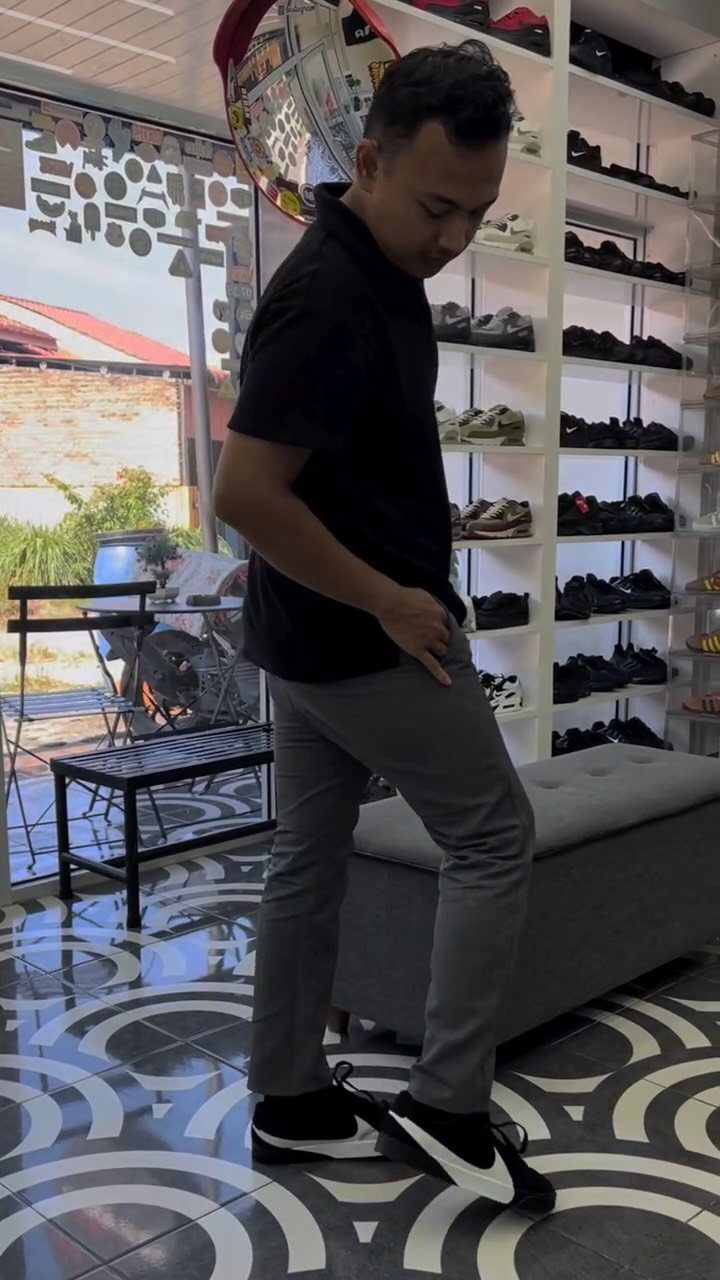 Man wearing black polo shirt, grey trousers, and black sneakers in a shoe store.