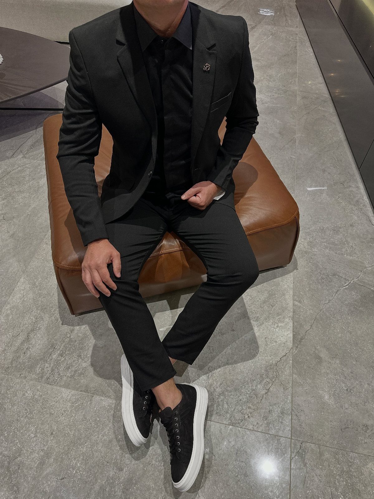 Man in a black suit wearing black sneakers with white soles sitting down.