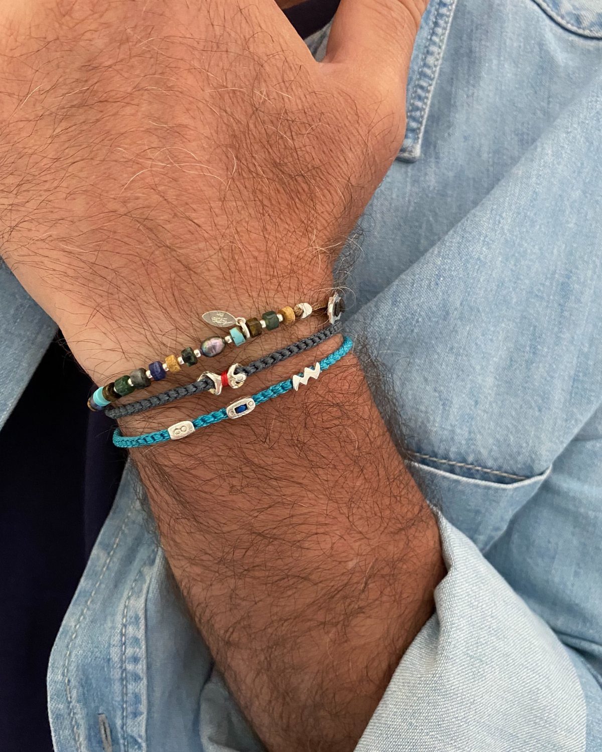 Man's wrist wearing three braided summer bracelets and a light blue denim shirt.