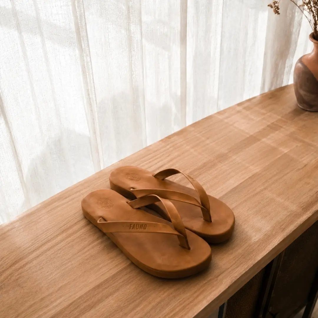 Brown leather flip-flops on a wooden table with white curtains in background