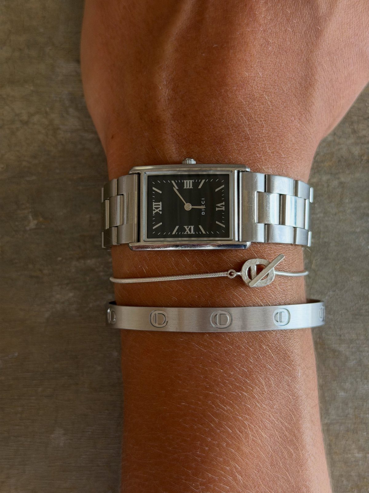 Tanned arm wearing a silver rectangular watch and two silver bracelets.