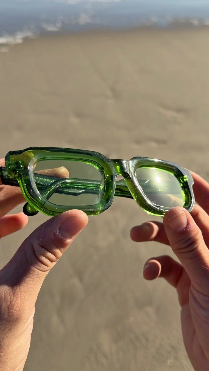 Translucent green sunglasses held by hands over a sandy beach and ocean.