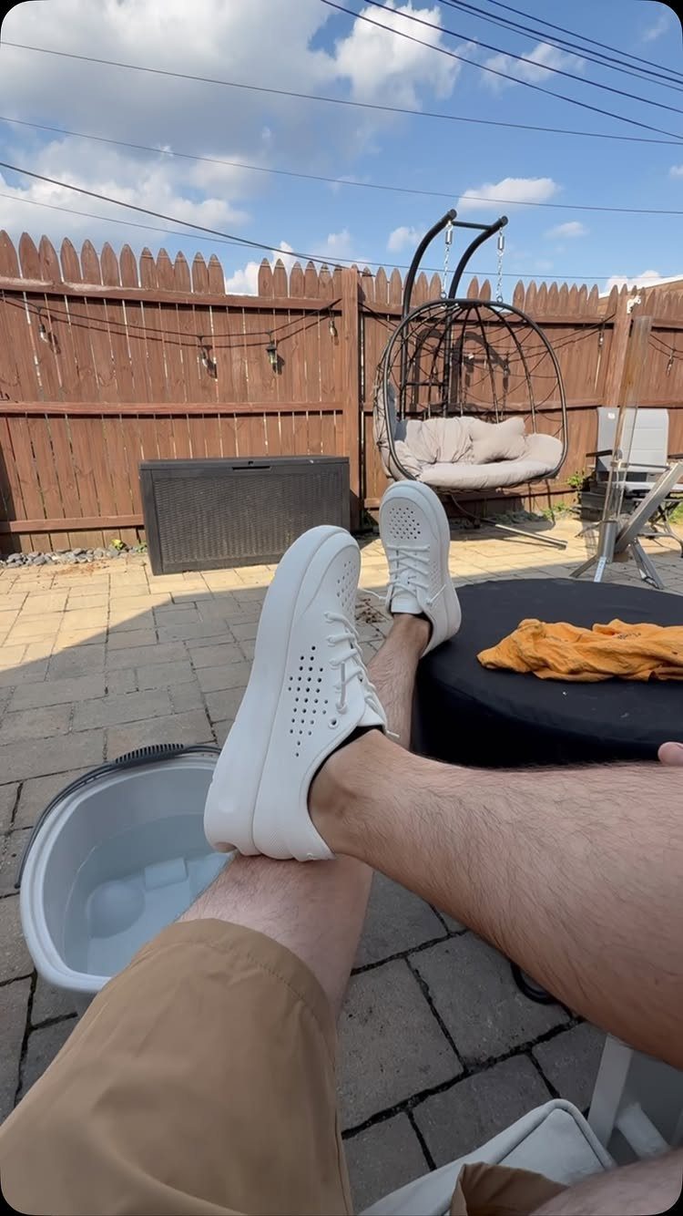 White perforated slip-on sneakers on a person's feet resting on a stone patio.