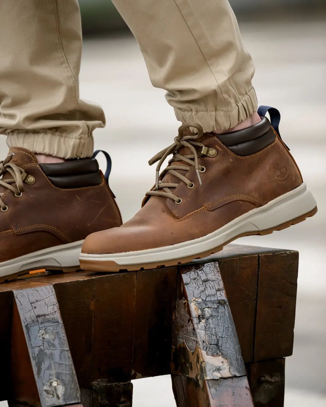 Brown leather ankle boots with light soles worn with tan trousers on wood.