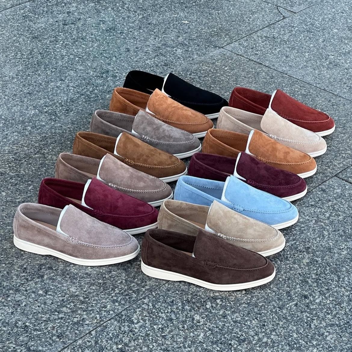 Various colored suede sneaker loafers with white soles arranged on grey stone pavement.