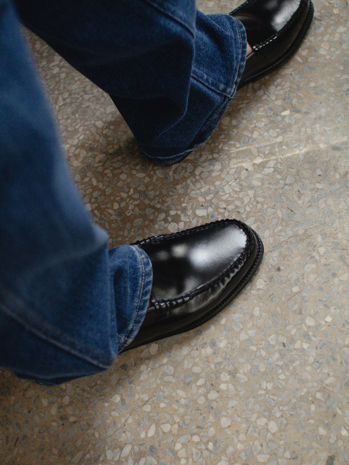 Black leather Venetian loafers worn with blue denim jeans on a stone floor.