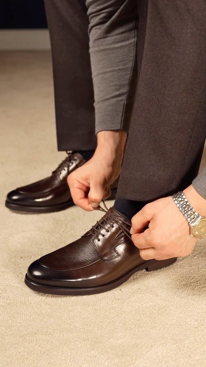 Dark brown leather derby shoes being tied by a man in grey trousers.
