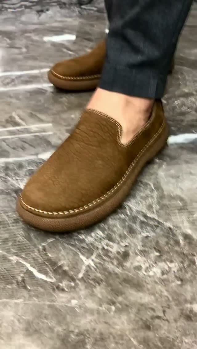 Brown leather slip-on loafers worn with dark grey trousers on marbled flooring.