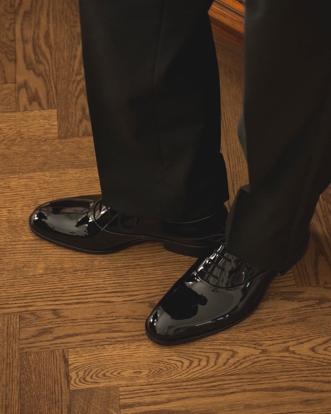 Shiny black patent leather tuxedo shoes worn with black dress pants on wood flooring