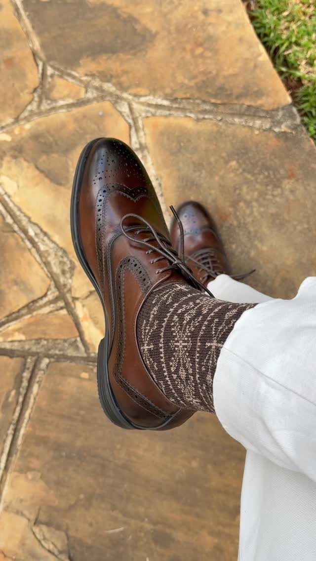 Brown leather wingtip shoes with patterned socks and white trousers on stone pavement.