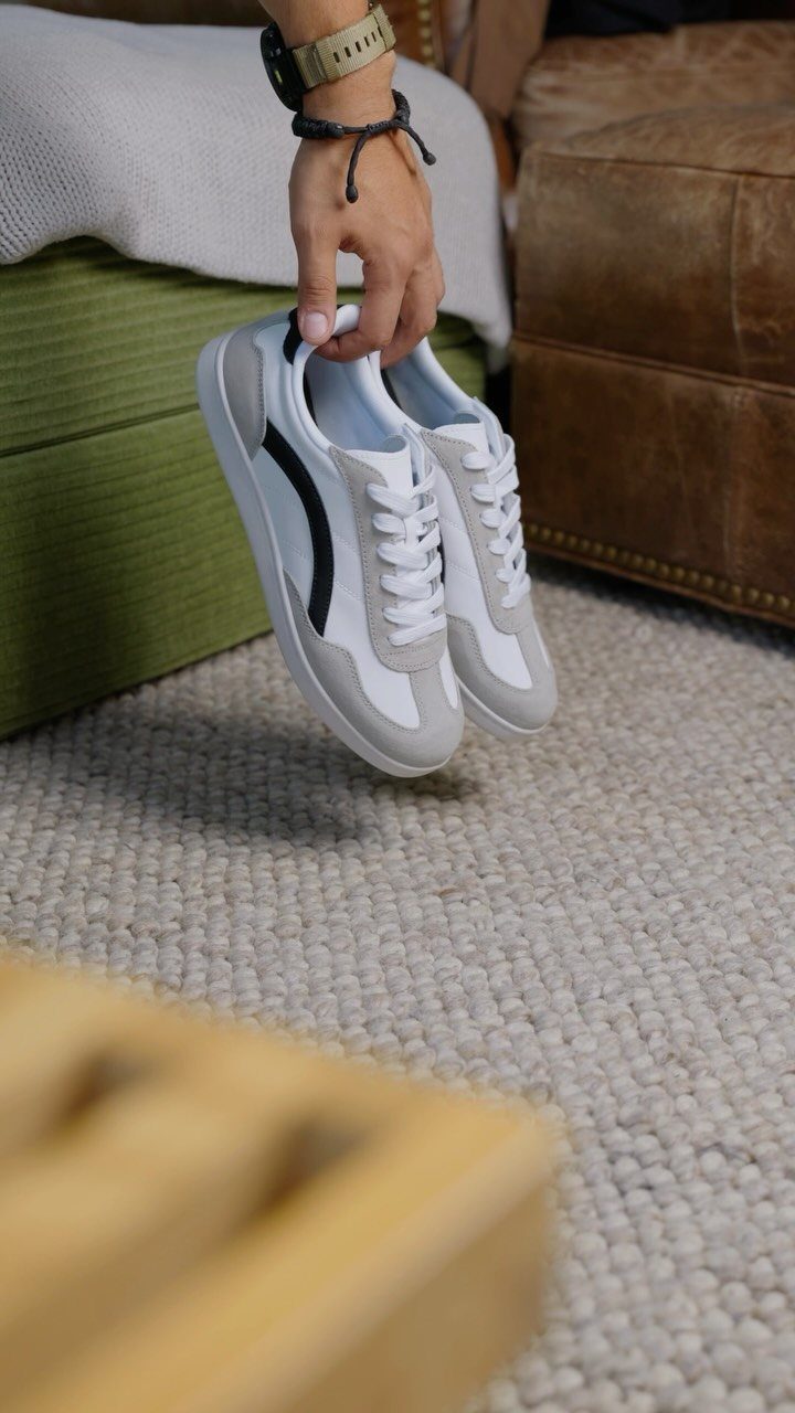 White sneakers with black stripes held by a hand over a textured rug