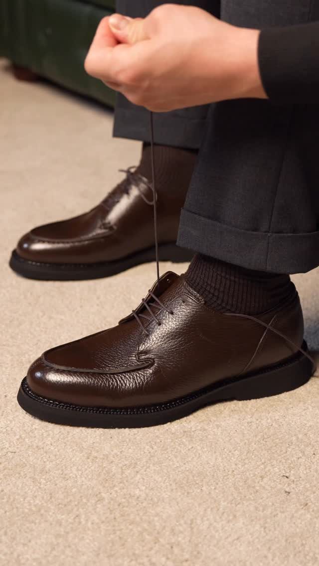 Dark brown leather derby shoes being tied by a person in grey trousers.
