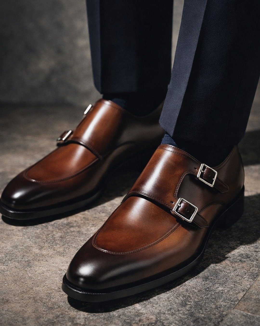Brown leather double monk strap shoes with silver buckles and navy trousers.