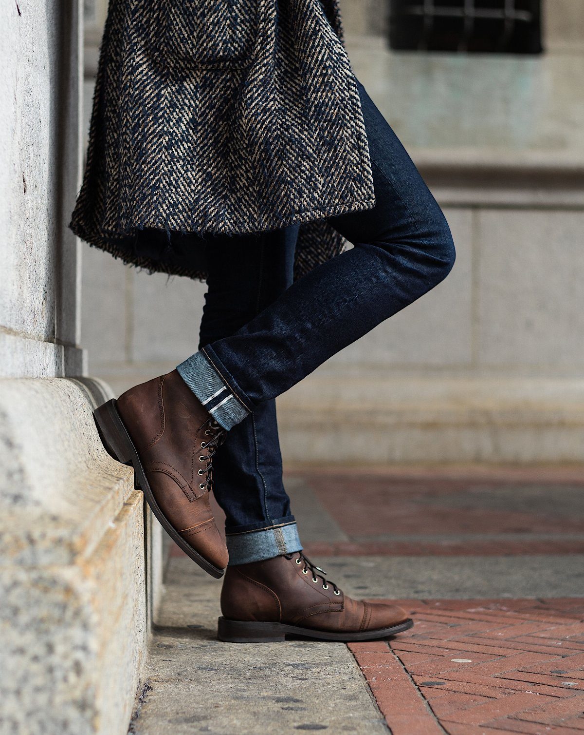Person wearing a herringbone wool coat, dark denim jeans, and brown leather boots.