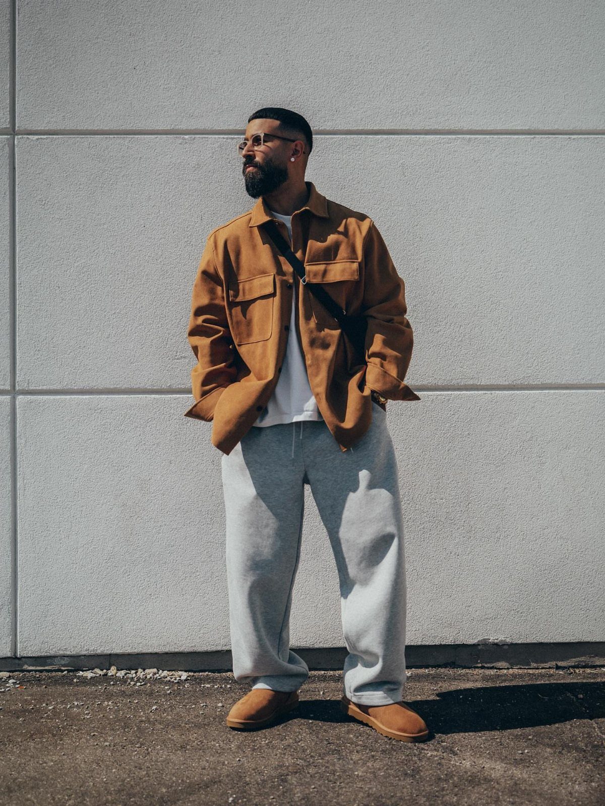 Man wearing brown shirt, grey sweatpants, and tan suede slippers against white wall