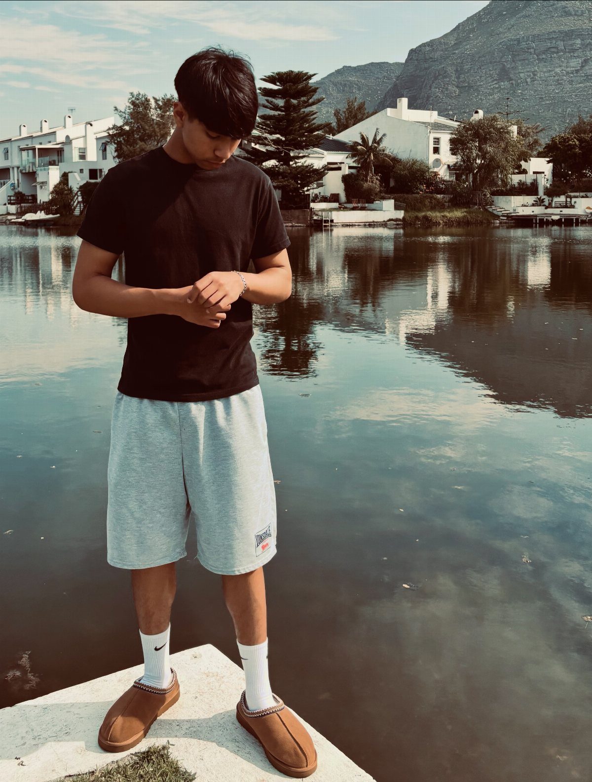 Person wearing black t-shirt, grey shorts, white socks, and tan Ugg slides by water.