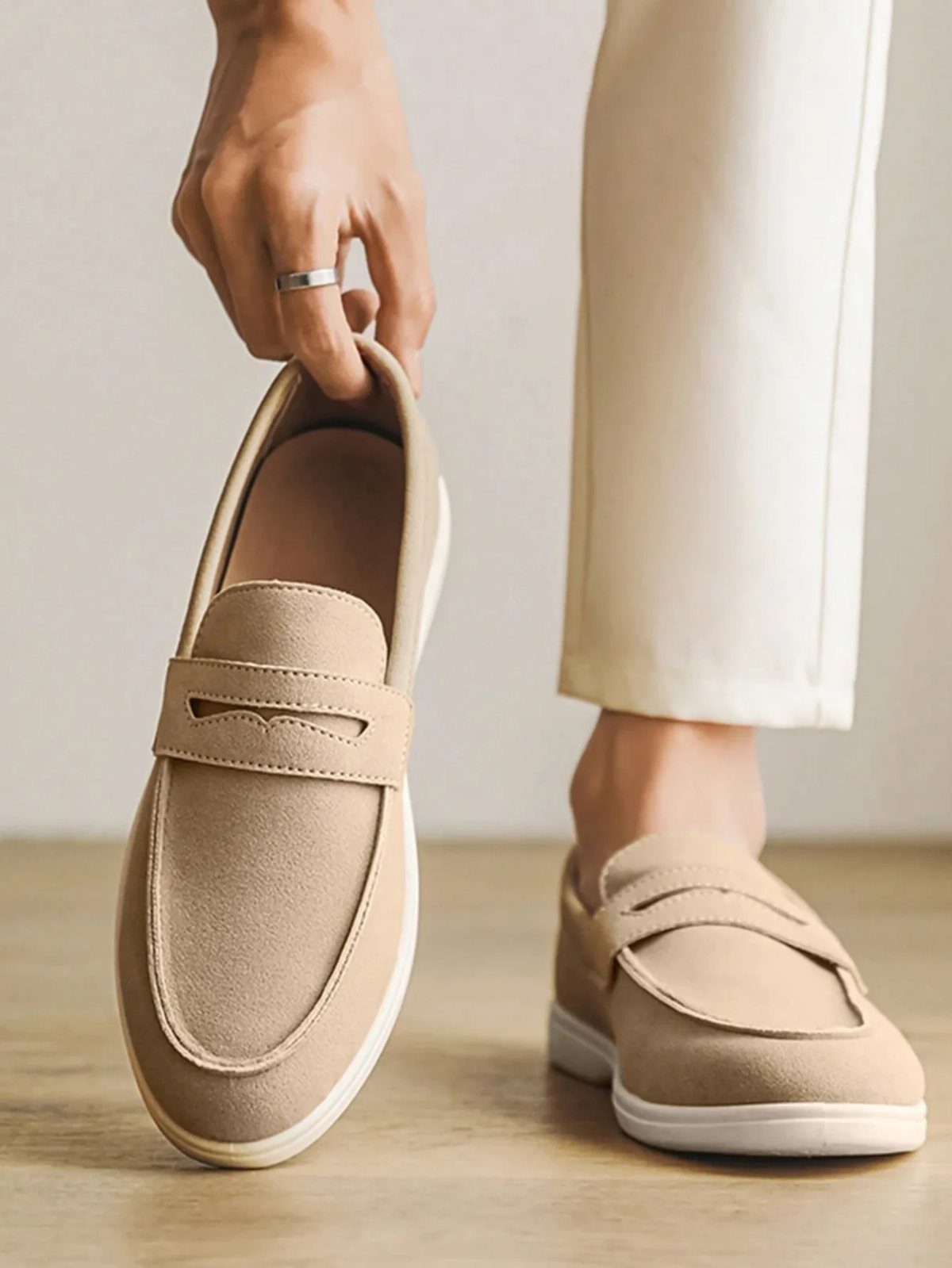 Beige suede loafers paired with white trousers on a light wooden floor.