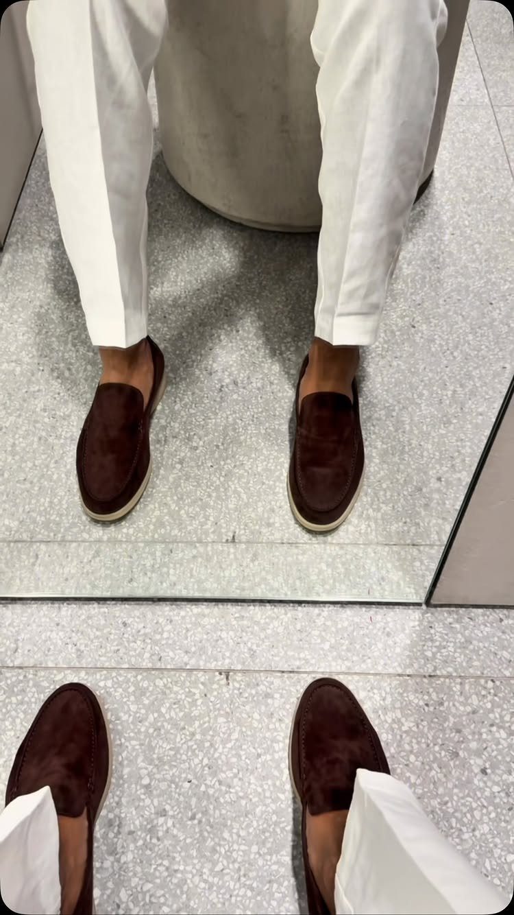 Dark brown suede loafers worn with white trousers on a grey speckled floor.