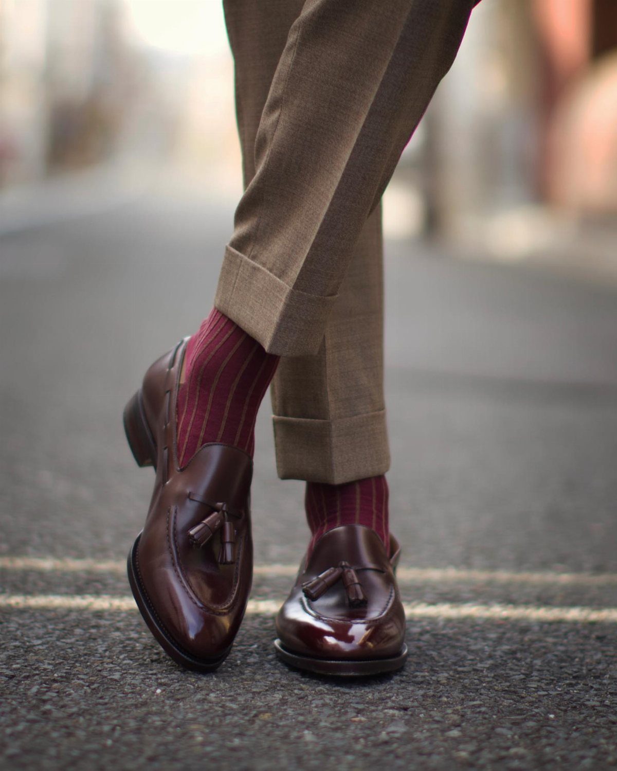 Dark brown tassel loafers and burgundy striped socks with brown trousers on asphalt.