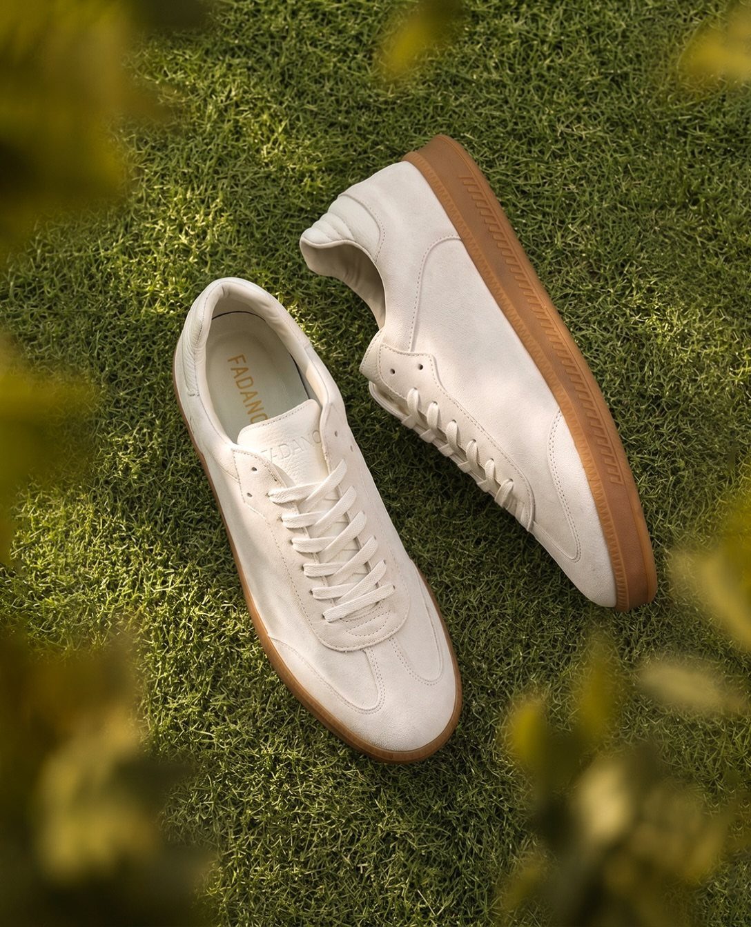 White leather dress sneakers with brown soles resting on a green grass lawn.