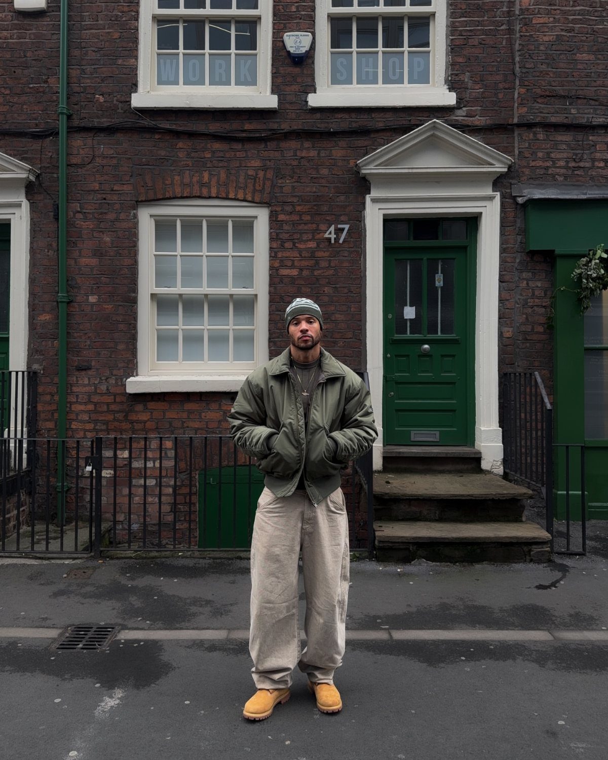 Man wearing olive jacket, beige trousers, and tan boots in front of brick house