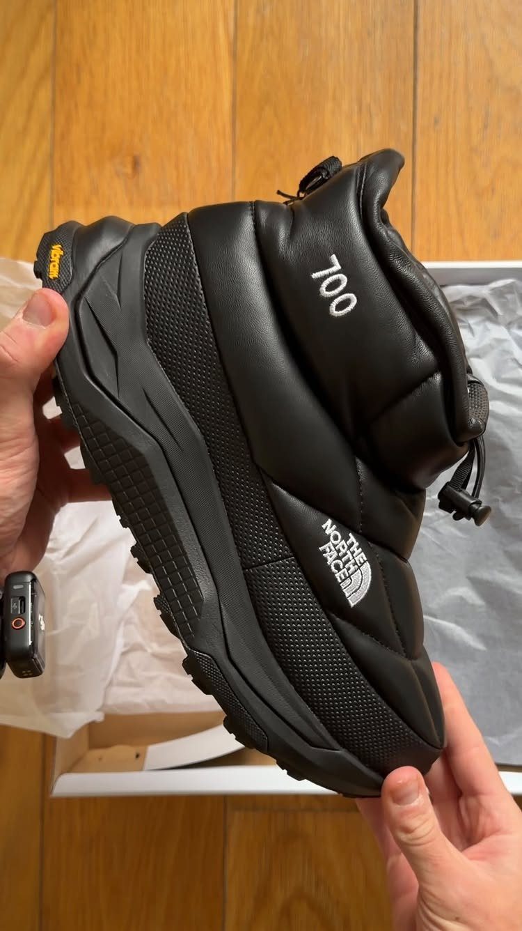 Black puffer winter boot held by hands over a white shoebox on wood.